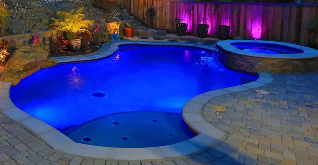 Luxurious backyard pool with vibrant blue LED lighting, spa, and paver deck at night with landscape lighting.