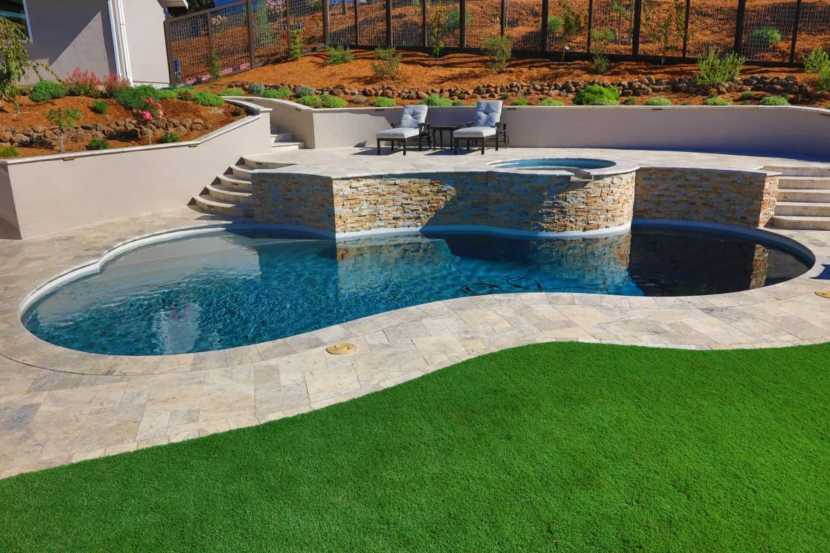 Custom freeform pool with a raised stacked stone spa, travertine deck and turf lawn, built by Royal Pools by Adams in Scotts Valley, CA